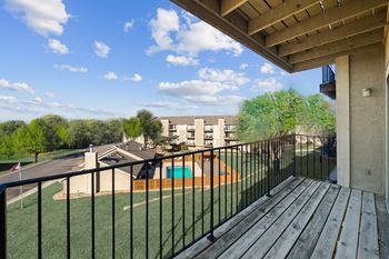 A balcony with a black railing and a wooden floor overlooks a green lawn and a building.